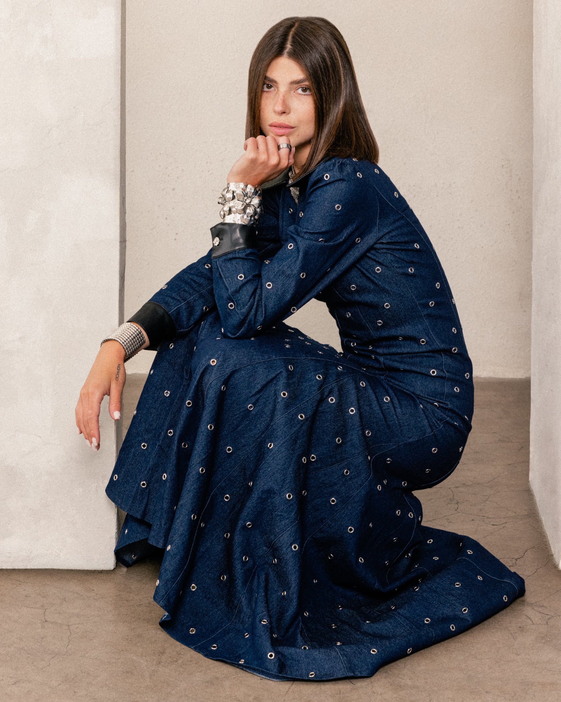 A woman modeling a dark navy blue long-sleeve dress with small white embroidered dots and black collar/cuff accents, posed against a light gray studio background.