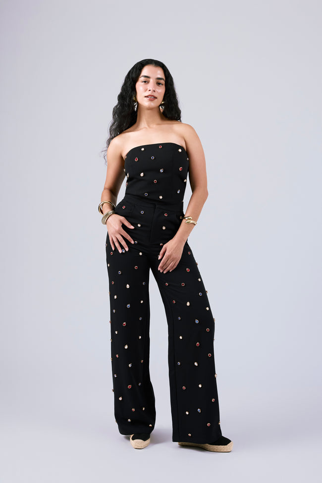 A woman modeling a black strapless jumpsuit with gold and rose gold metallic dot embellishments, set against a solid light gray studio background.