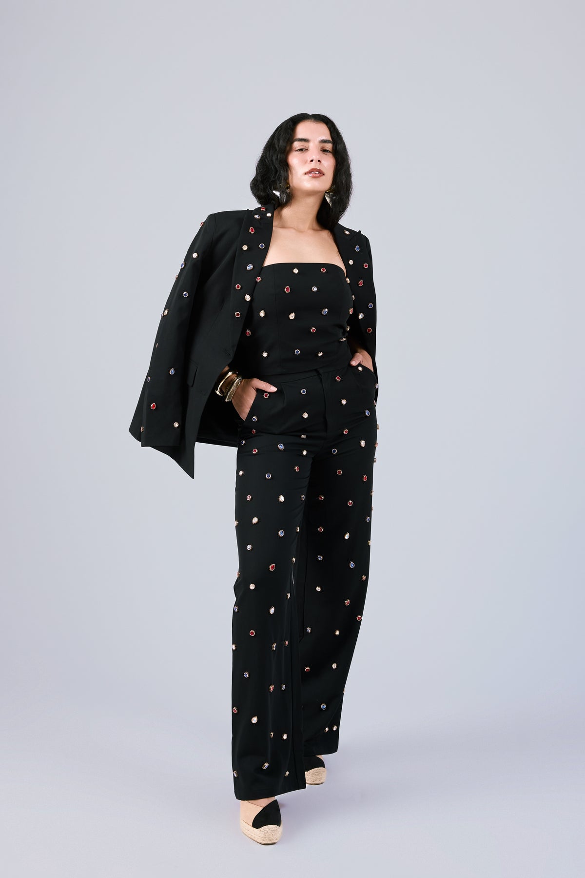 A model wearing a black three-piece jumpsuit set with colorful embroidered dots, featuring a wide-leg trouser, a strapless top, and a matching blazer draped over one shoulder, set against a solid light gray background.