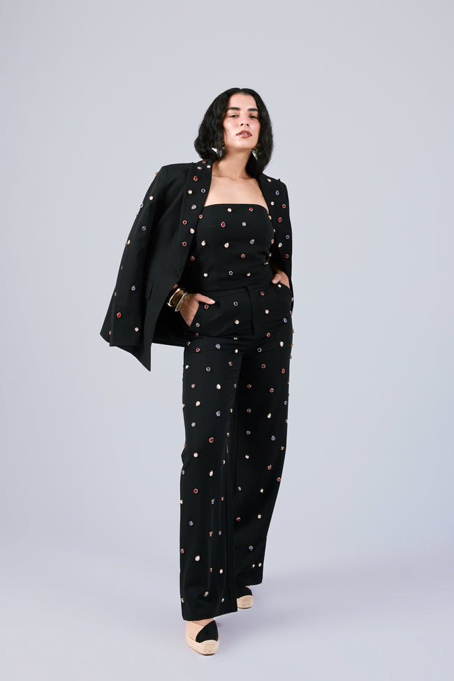 A model wearing a black three-piece jumpsuit set with colorful embroidered dots, featuring a wide-leg trouser, a strapless top, and a matching blazer draped over one shoulder, set against a solid light gray background.