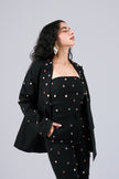 A woman modeling a black three-piece outfit with colorful embroidered dots, consisting of a strapless top, matching blazer, and trousers, set against a solid gray background.