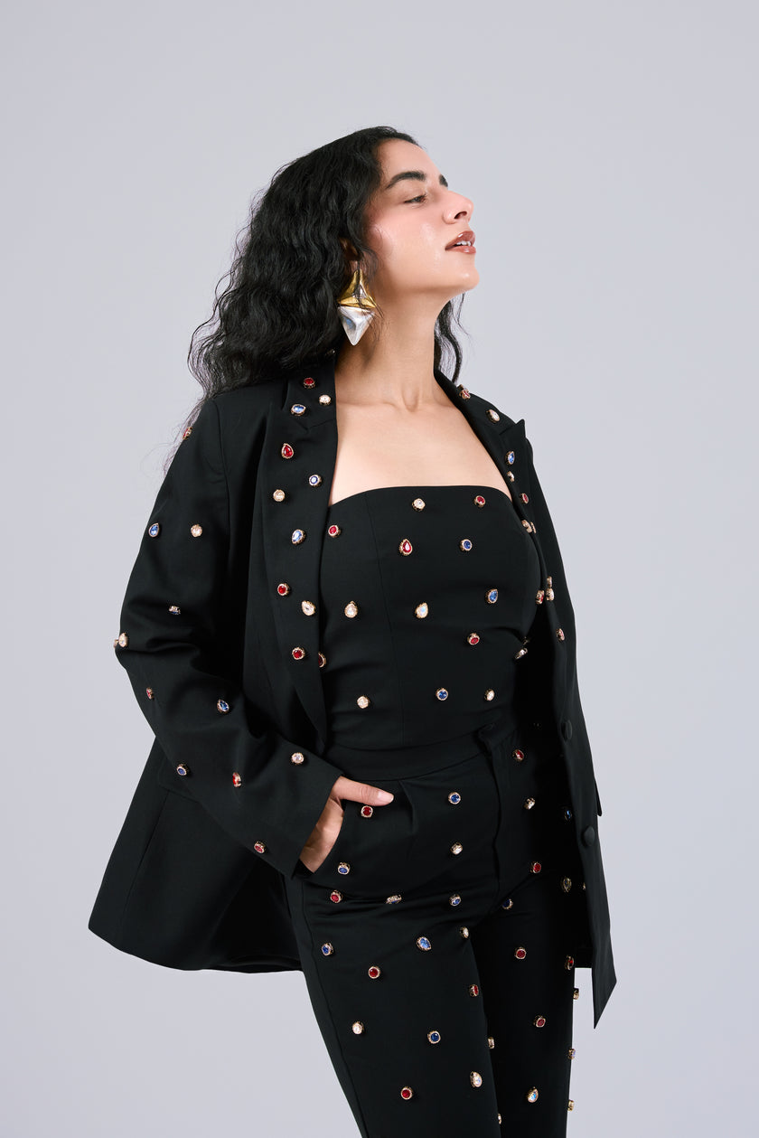 A woman modeling a black three-piece outfit with colorful embroidered dots, consisting of a strapless top, matching blazer, and trousers, set against a solid gray background.