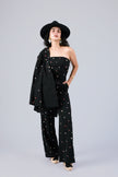 A model wearing a black one-shoulder strapless top, matching wide-leg trousers, and a black blazer draped over one shoulder, styled with a black wide-brim hat, gold earrings, and beige heels against a light gray studio background.