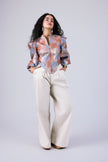 A woman modeling a long-sleeve button-up shirt with a colorful abstract pattern in orange, blue, and white, paired with wide-leg cream trousers, against a light gray studio background.