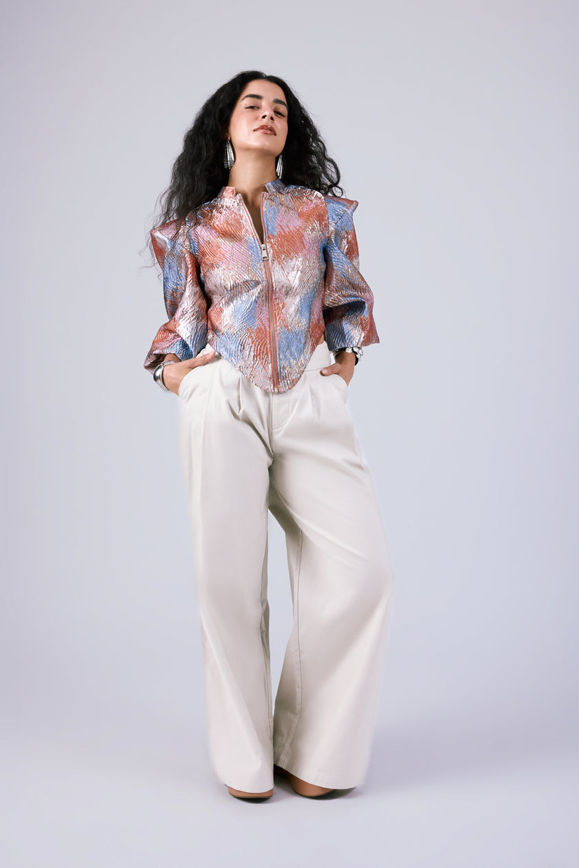 A woman modeling a long-sleeve button-up shirt with a colorful abstract pattern in orange, blue, and white, paired with wide-leg cream trousers, against a light gray studio background.