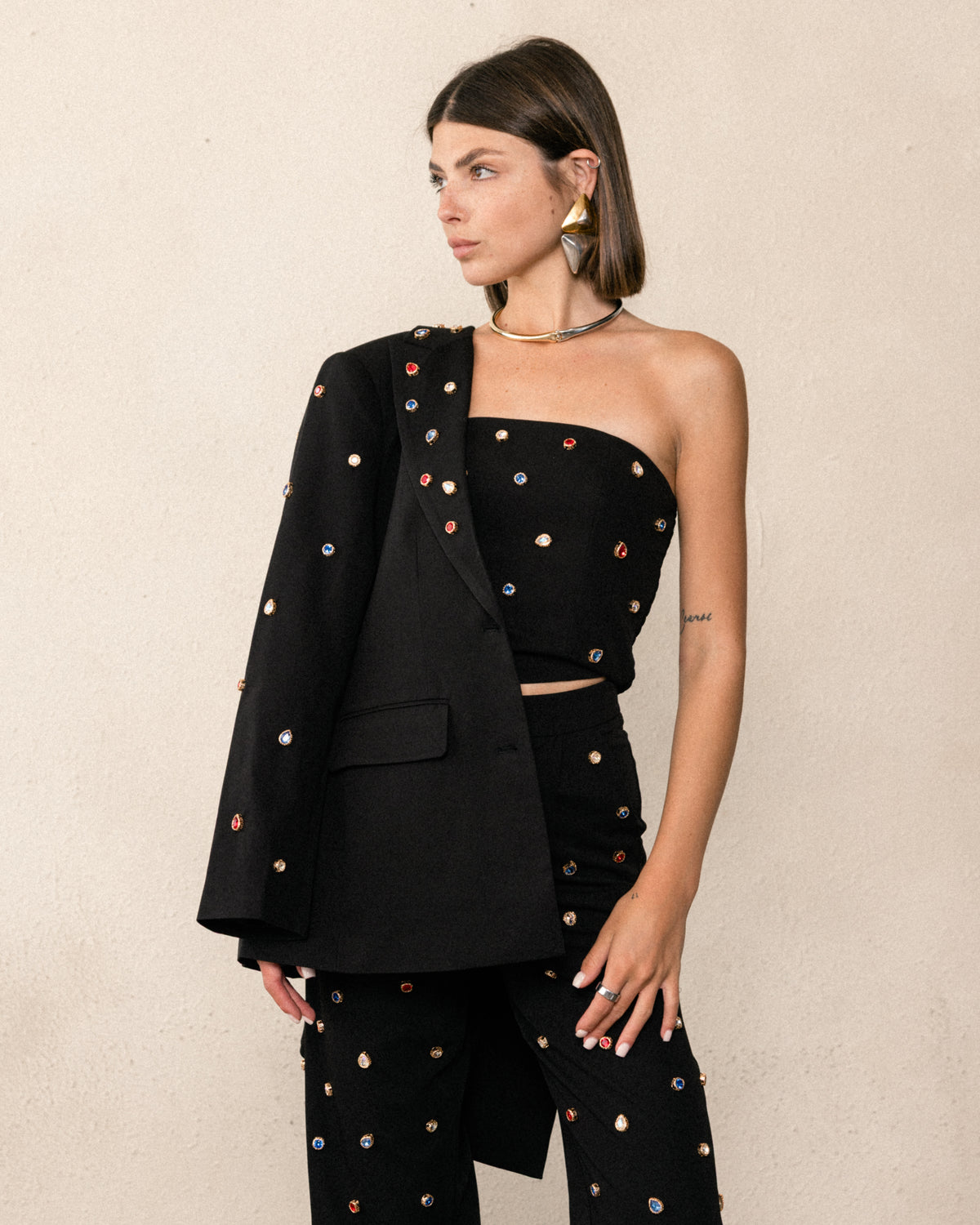 A model wearing a black three-piece outfit consisting of a strapless tube top, a matching blazer draped over one shoulder, and trousers, all embellished with colorful embroidered dots, set against a textured beige wall.