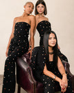 Three women modeling matching black strapless jumpsuits with colorful embroidered dots, posed together on a dark purple leather armchair against a neutral beige wall.