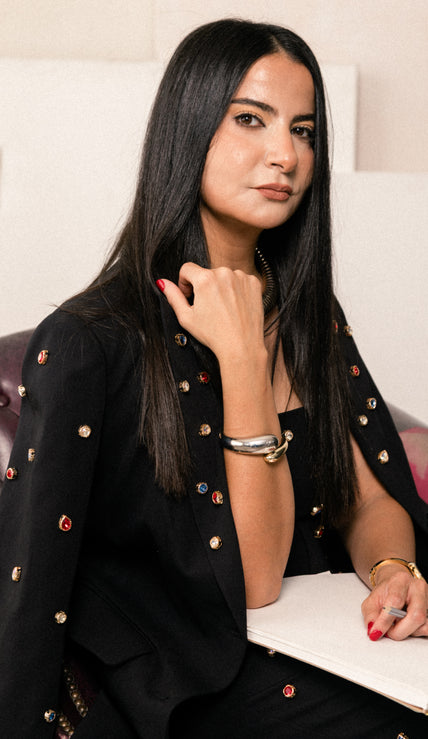 A woman with long dark hair sits in a purple leather chair, wearing a black blazer adorned with colorful rhinestone embellishments. She is holding a white notebook on her lap, with a neutral indoor background.