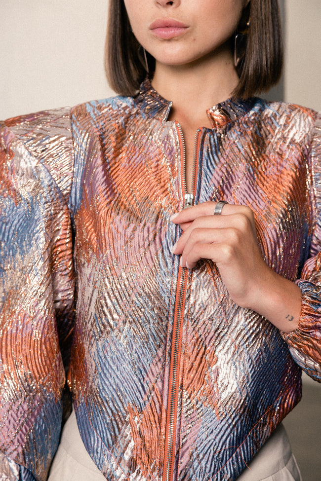 A close-up of a model wearing a metallic, textured zip-up jacket with a shimmering pattern of copper, silver, and blue.