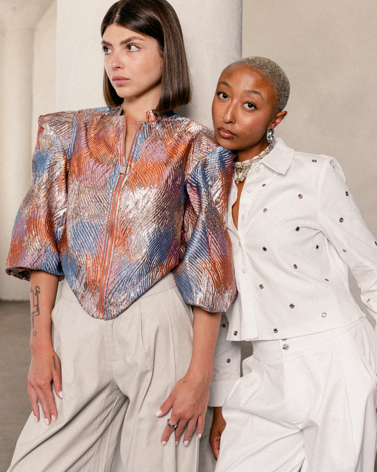 Two models posing in a studio setting. The model on the left wears a metallic, textured cropped jacket with orange, blue, and silver tones, paired with light beige trousers.