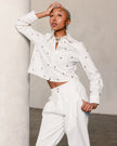 A model with short blonde hair poses in a white cropped long-sleeve button-up shirt and matching white trousers, both decorated with small metallic studs, set against a textured light gray wall.