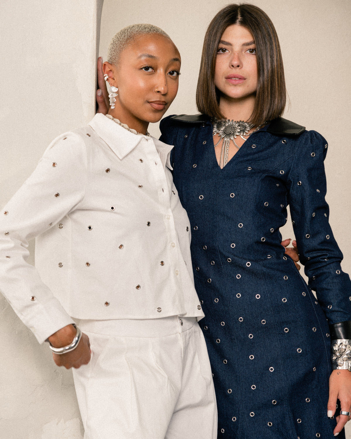Two fashion models posing together. The model on the left wears a white collared jacket and matching trousers, both adorned with small metallic stud embellishments.
